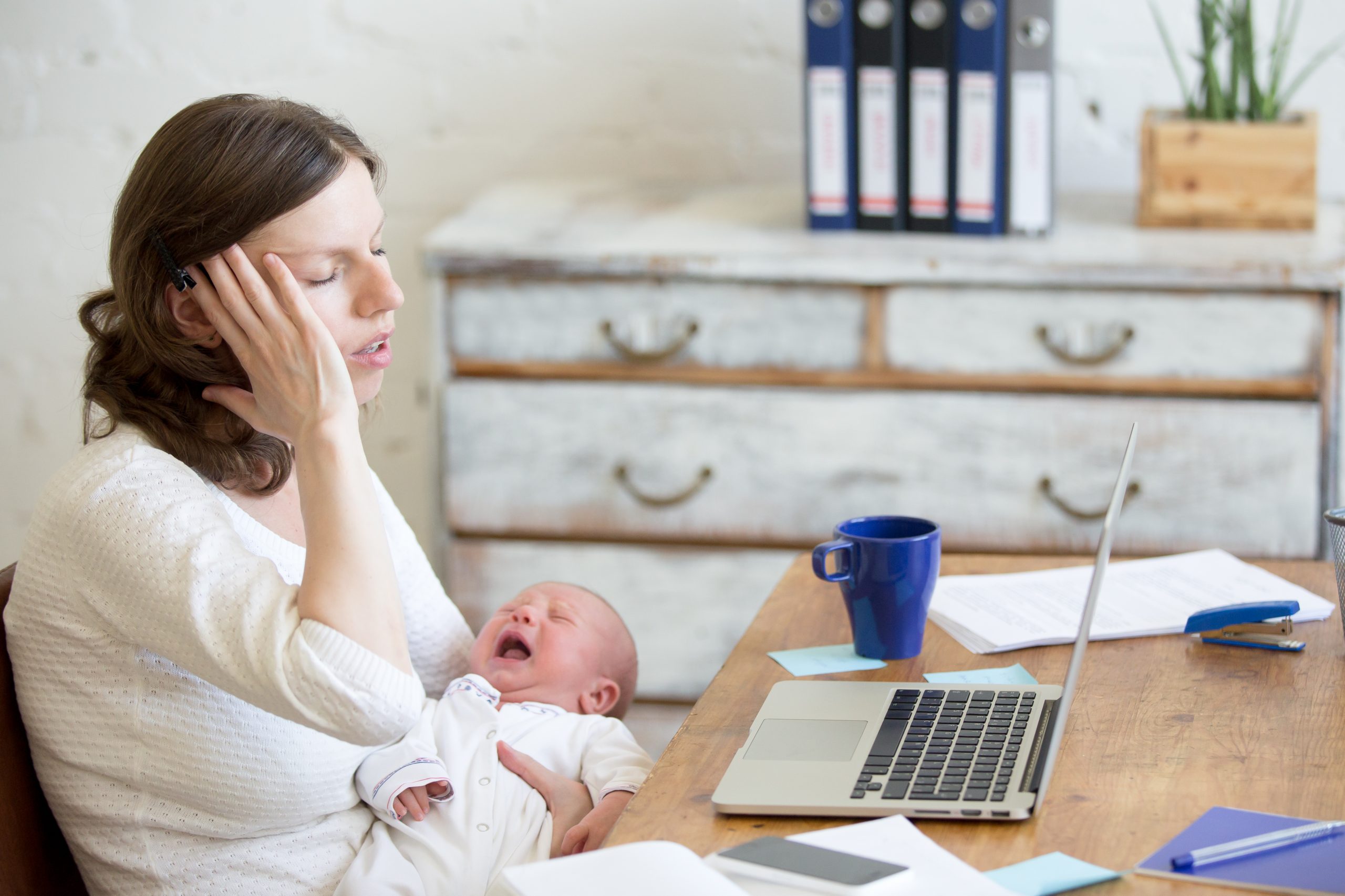 young,businesswoman,holding,newborn,crying,babe,while,sitting,with,laptop